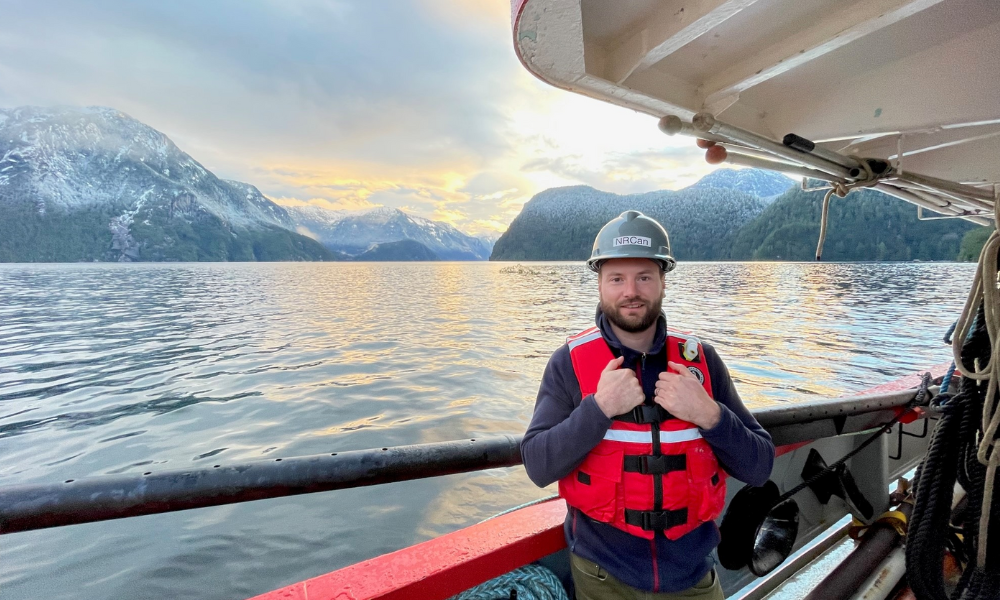 A scientist facing the camera and standing on the deck of a boat with the ocean and mountains in the background.