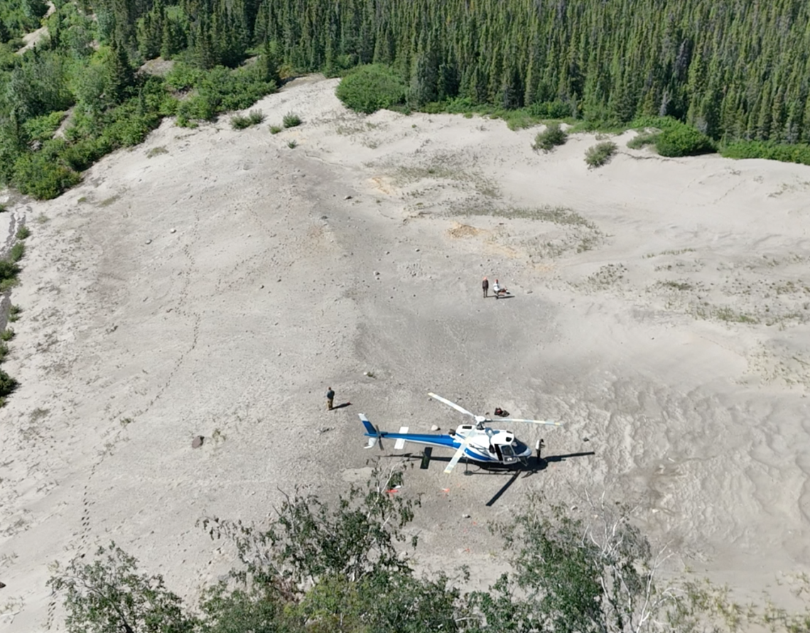 Aerial view of a helicopter resting on a sand dune with a few people in the surrounding area studying the surface. 