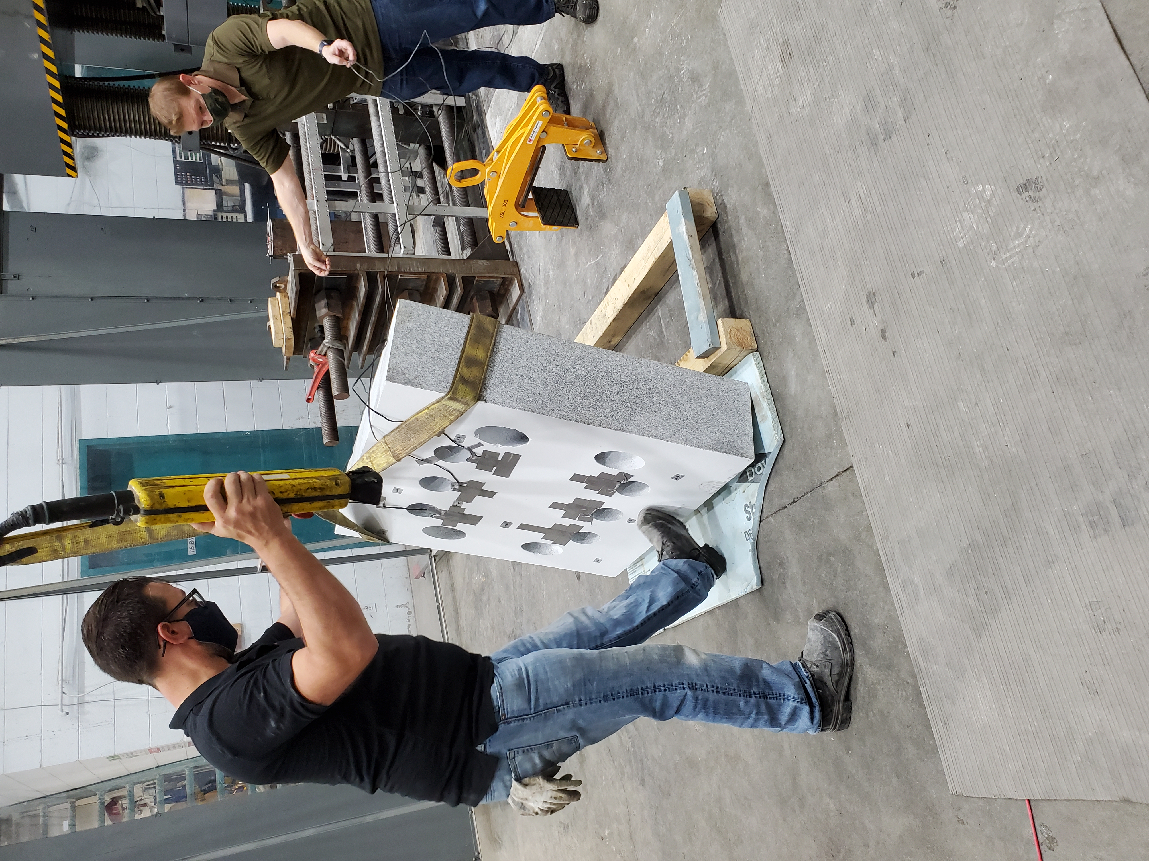 A large industrial testing machine with vertical hydraulic columns is positioned behind a transparent protective barrier on wheels.