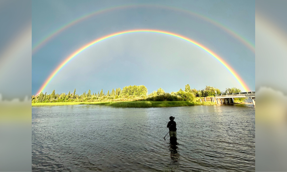 Silhouette of a man fishing on a wide river with a double rainbow in the background.
