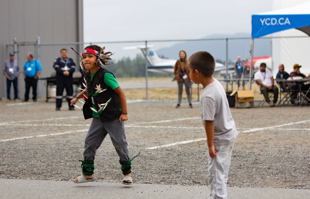 Two children dance on a tarmac at an airport, while a group of people watch.