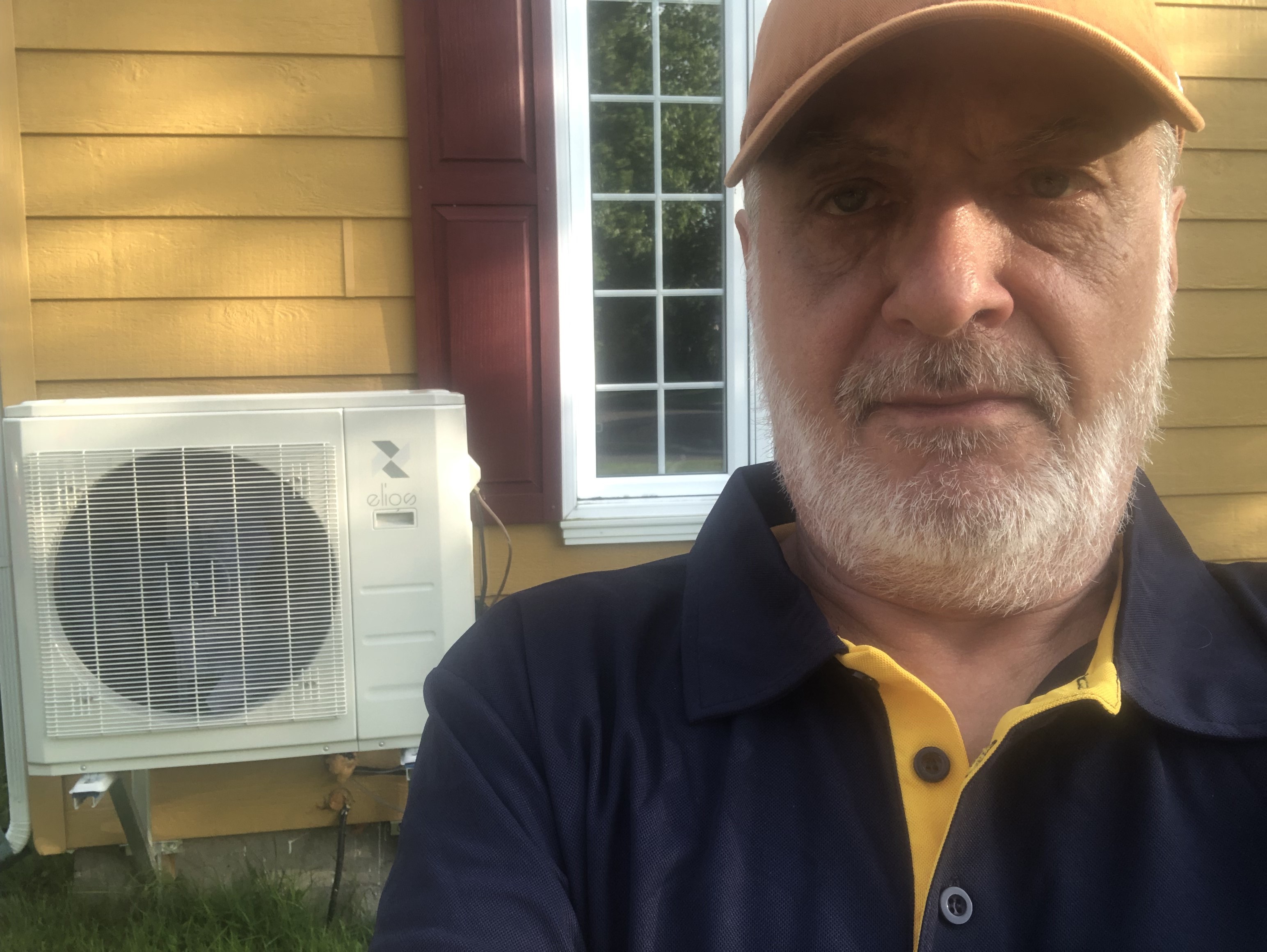 A man takes a selfie in front of his house, with a heat pump in the background.