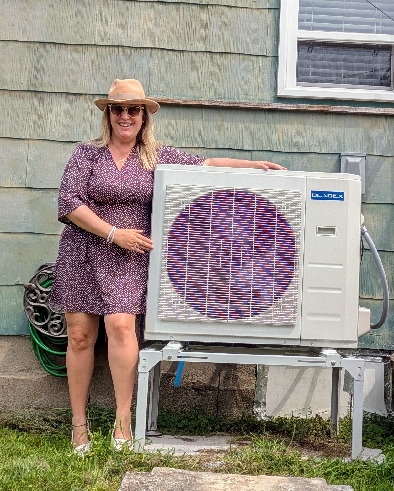 Another smiling woman rests her arm on a heat pump on a metal stand next to her home