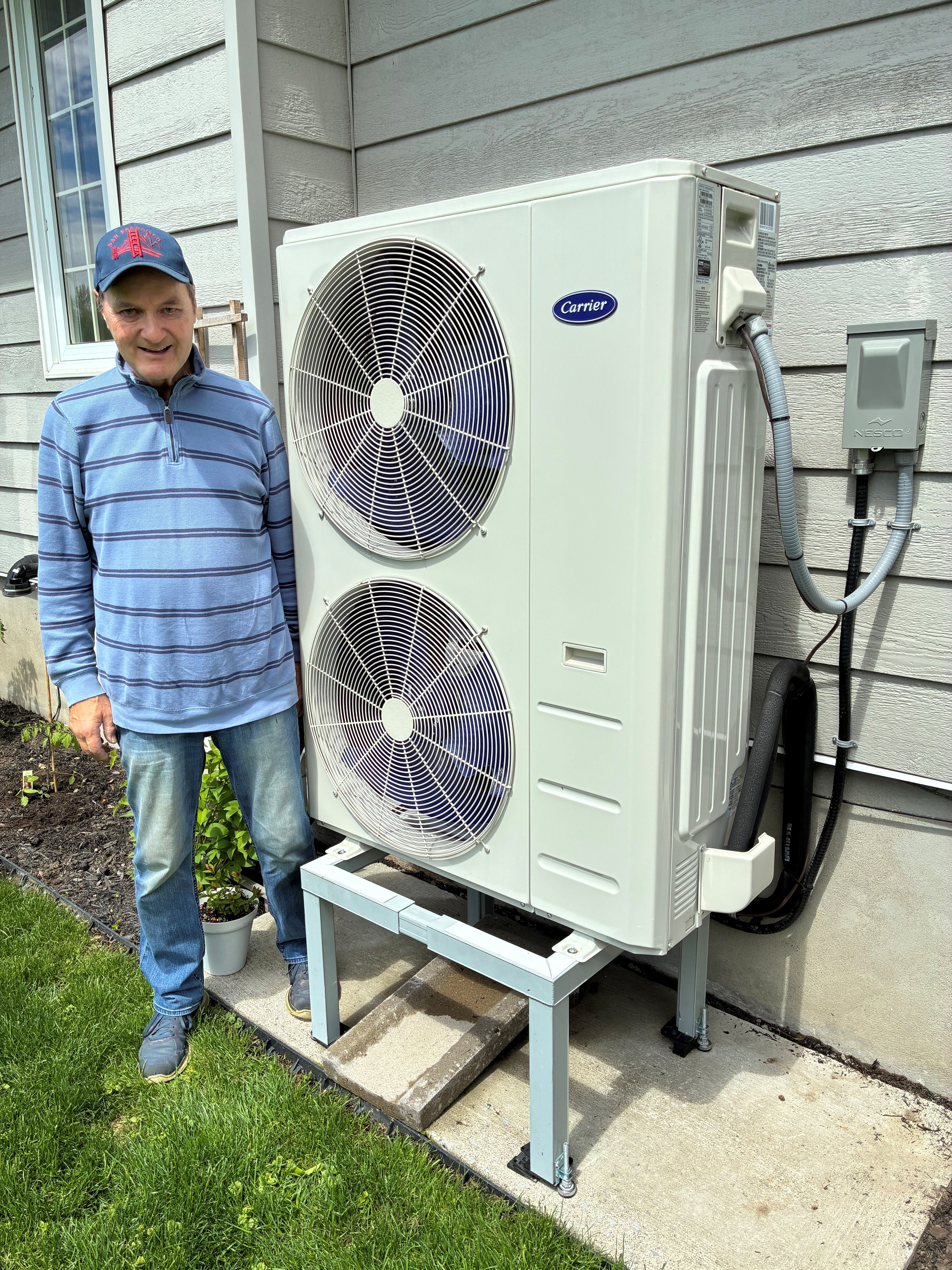 A smiling man near a heat pump on a metal stand next to his home