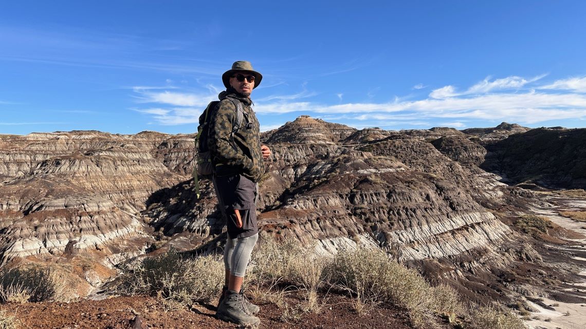 Researcher wearing hiking gear stands on the edge of a dry mountainous landscape with a bright blue sky behind him. 