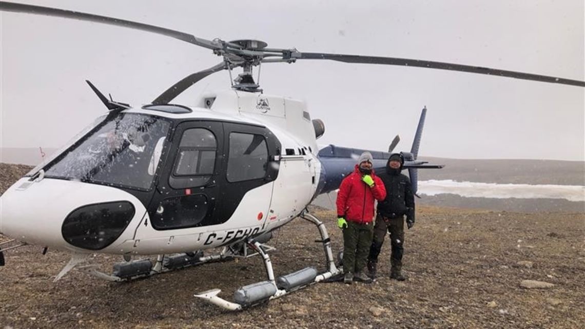 Two researchers in cold weather gear standing in front of a helicopter on a flat rocky landscape.  