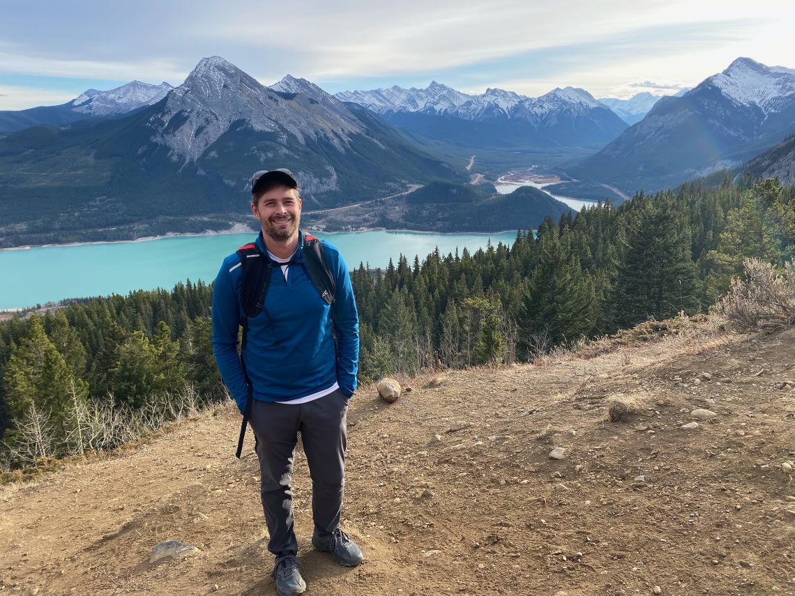 A man standing on a sloping hillside, with a forest, river and mountain range in the background. 