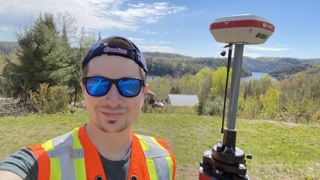 Researcher in a safety vest and wearing sunglasses holds a piece of equipment standing in a field with a river in the background.