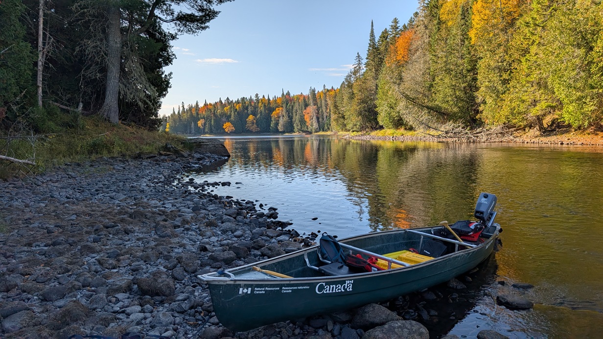 A small green Government of Canada canoe is pulled up along a rocky riverbank, carrying fieldwork gear in a remote setting.