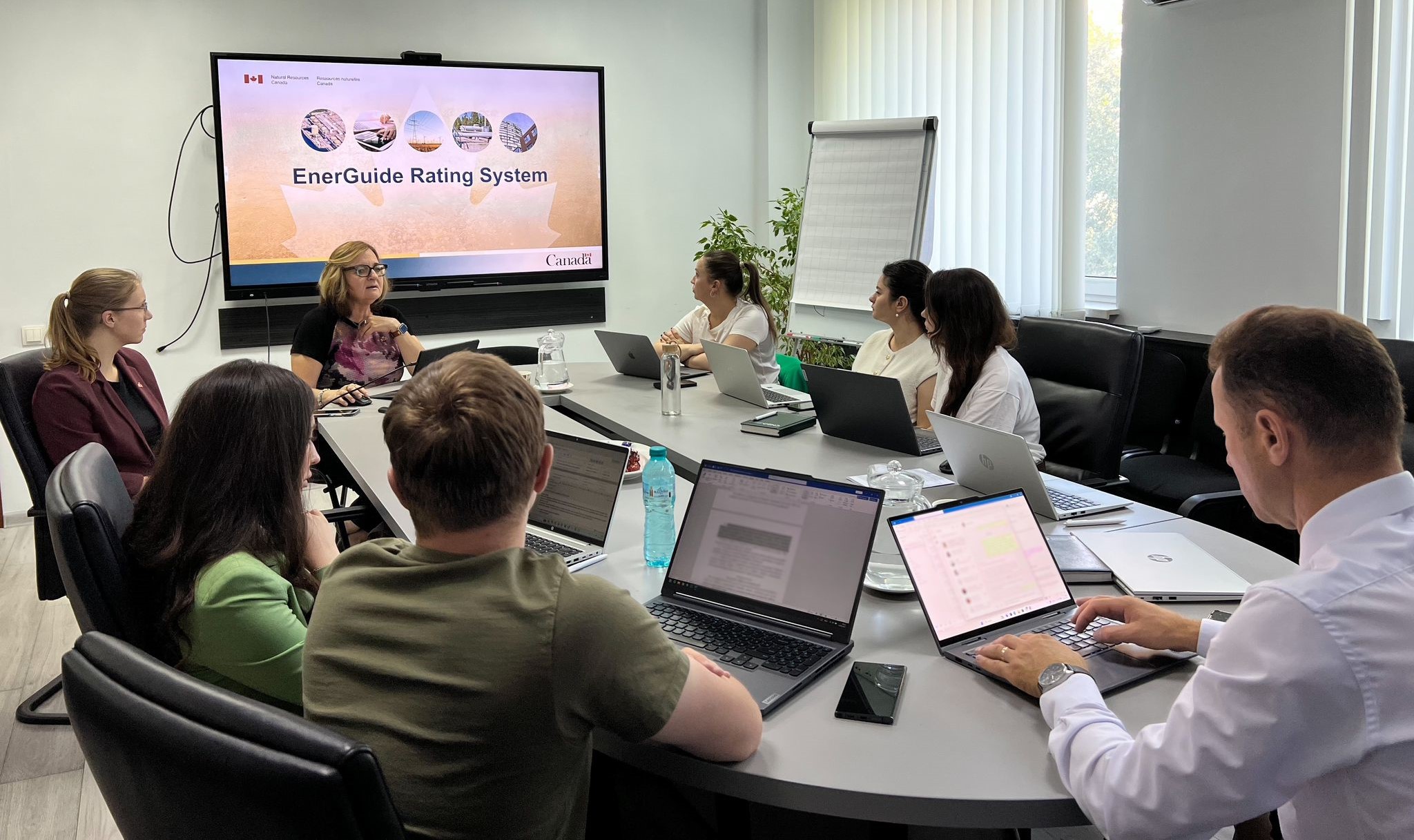 A business meeting with eight people seated around a conference table, focusing on a presentation displayed on a large screen.