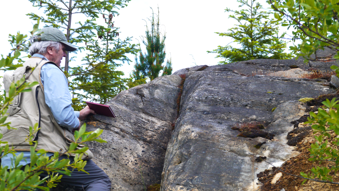 Geologist in a beige vest examines a rock outcrop, holding a notebook against its surface.