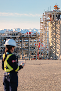 A woman with a clipboard and hard hat looking at a liquid natural gas storage tank.