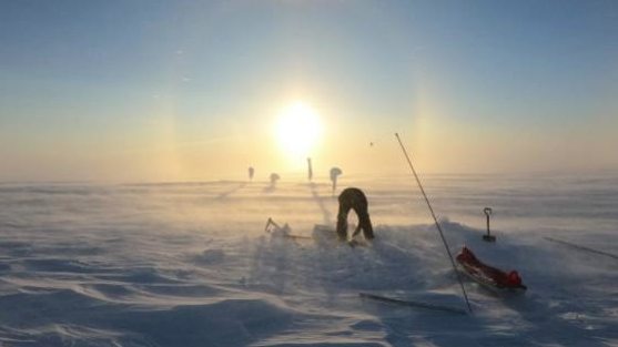 A researcher works in a snow-covered ice field in low sunlight. Blowing snow moves across the surface, with scientific equipment and a sled visible nearby.