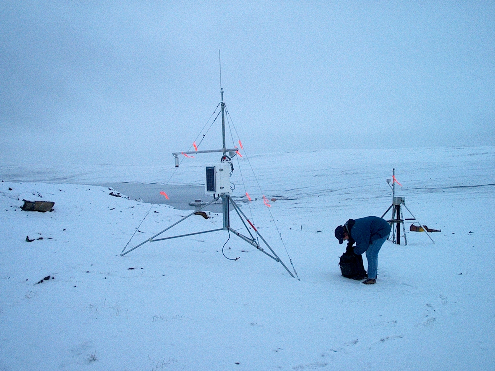 A researcher stands beside permafrost monitoring equipment on a flat, snow-covered landscape in Alert, Nunavut. Tripods, cables and instruments are positioned at the monitoring site.